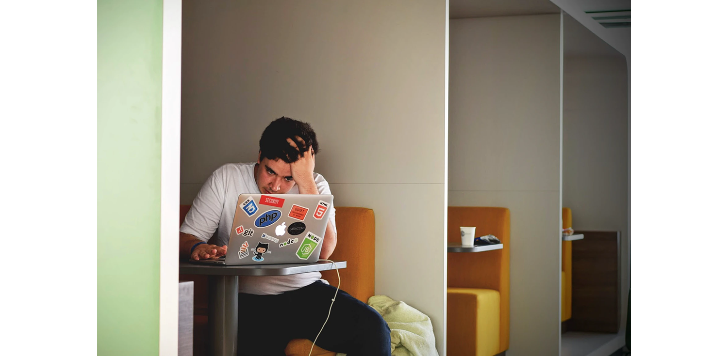 A man in a white t-shirt sitting in a booth while struggling with something on his laptop